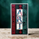 A vertical decorative card or art print with the word “ART” spelled out in large patterned letters, set against red, black, and teal abstract panels, photographed standing on a rustic wooden surface with a dark teal blurred background.