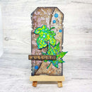 A handmade tombstone-style autumn card or plaque displayed on a small wooden easel. The card features distressed brown and beige textures, blue circular embellishments, and layered bright green and yellow maple leaves.