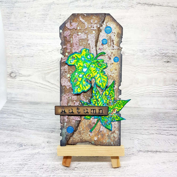 A handmade tombstone-style autumn card or plaque displayed on a small wooden easel. The card features distressed brown and beige textures, blue circular embellishments, and layered bright green and yellow maple leaves.