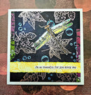 A handmade square greeting card featuring a dragonfly design over white leaf outlines on a dark background, with a bright yellow banner at the bottom that reads, 'I'm so thankful for you every day.' The card is photographed flat on a textured brown and orange patterned surface.