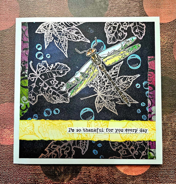A handmade square greeting card featuring a dragonfly design over white leaf outlines on a dark background, with a bright yellow banner at the bottom that reads, 'I'm so thankful for you every day.' The card is photographed flat on a textured brown and orange patterned surface.