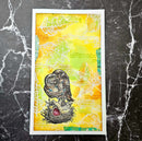 A framed art print featuring a detailed illustration of an owl perched on a nest, set against a vibrant yellow and green watercolor background with faint script and leaf patterns. The print is displayed on a dark gray marble surface.