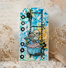 A decorative handmade paper tag or card featuring a colorful abstract blue and yellow background with black ink splatters, a film strip graphic, and a central illustration of a bird perched on a cupcake. The tag is displayed on a lace surface with dried flowers in the background.