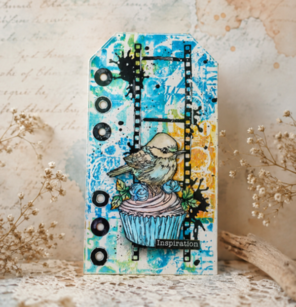 A decorative handmade paper tag or card featuring a colorful abstract blue and yellow background with black ink splatters, a film strip graphic, and a central illustration of a bird perched on a cupcake. The tag is displayed on a lace surface with dried flowers in the background.