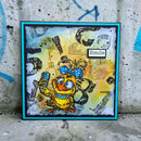 A square handmade art card featuring a cartoon yellow duck character with a blue polka-dot bow, set against a distressed yellow and gray background with the word 'Smile' in a small label. The card is framed in turquoise and is photographed leaning against a concrete wall with graffiti.