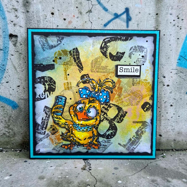 A square handmade art card featuring a cartoon yellow duck character with a blue polka-dot bow, set against a distressed yellow and gray background with the word 'Smile' in a small label. The card is framed in turquoise and is photographed leaning against a concrete wall with graffiti.