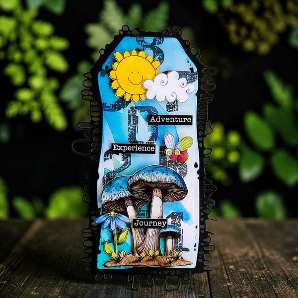 A decorative rectangular card or plaque with a black scalloped border, featuring a whimsical illustrated design of blue mushrooms, a smiling yellow sun, a white cloud, a bee, and the words 'Adventure', 'Experience', and 'Journey'. The item is photographed standing upright on a dark wooden surface with a blurred green garden background.