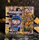 A handmade autumn-themed card featuring a cute cartoon girl in a blue beanie and sweater holding a pink cup, next to a 3D metallic oak leaf. The card has a distressed, grunge background with the text 'Fun falling for cozy' and is displayed on a rustic wooden surface with scattered leaves.