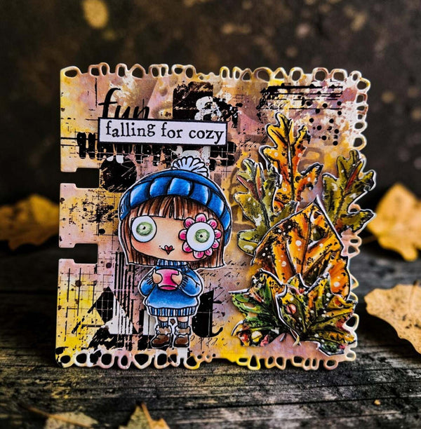 A handmade autumn-themed card featuring a cute cartoon girl in a blue beanie and sweater holding a pink cup, next to a 3D metallic oak leaf. The card has a distressed, grunge background with the text 'Fun falling for cozy' and is displayed on a rustic wooden surface with scattered leaves.