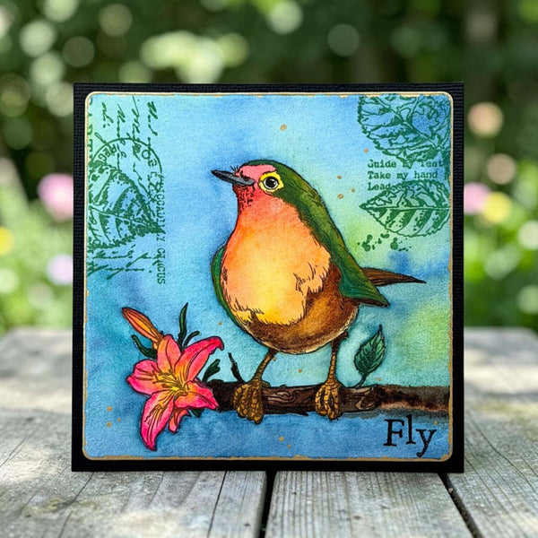 A square art card featuring a colorful illustration of a robin perched on a branch with a pink flower, set against a blue background with decorative text and leaf motifs. The card is displayed on a wooden surface with a blurred green garden background.