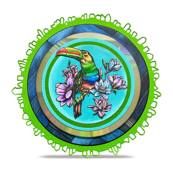 A round decorative plate featuring a colorful illustration of a toucan perched among pink flowers on a turquoise background, surrounded by concentric blue and gold rings and a bright green scalloped outer rim, shown on a white background.