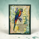A handmade greeting card featuring a colorful parrot perched among purple flowers, set against a vintage-style book page background with torn paper edges and inspirational text. The card is displayed on a light teal background with subtle watercolor splatters.
