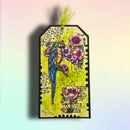 A rectangular decorative tag with scalloped edges, featuring a colorful parrot perched among pink flowers on a yellow-green textured background with black script text. A lime green tassel is attached at the top, and the tag is photographed against a soft pastel gradient background.