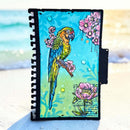 A spiral-bound notebook or journal standing upright on a sandy beach with the ocean blurred in the background. The cover features a colorful illustration of a green and yellow parrot perched among pink flowers on a bright turquoise and light green background.