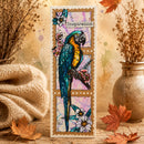 A vertical handmade greeting card featuring a detailed illustration of a blue and yellow macaw parrot perched among pink flowers, styled on a rustic wooden surface with autumn leaves, a knitted blanket, and dried flowers in a ceramic vase.
