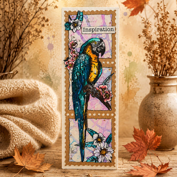 A vertical handmade greeting card featuring a detailed illustration of a blue and yellow macaw parrot perched among pink flowers, styled on a rustic wooden surface with autumn leaves, a knitted blanket, and dried flowers in a ceramic vase.