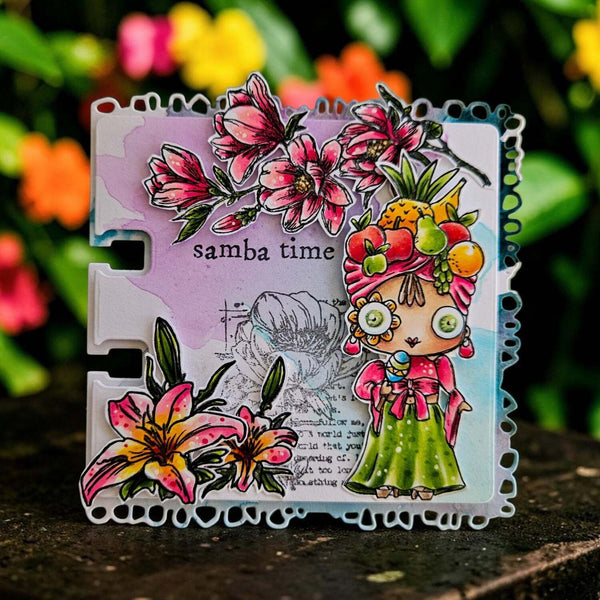 A colorful handmade art card with a scalloped white border, featuring illustrated flowers, a stylized woman in a green dress, and the text 'samba time', photographed outdoors against a blurred background of green foliage and flowers.