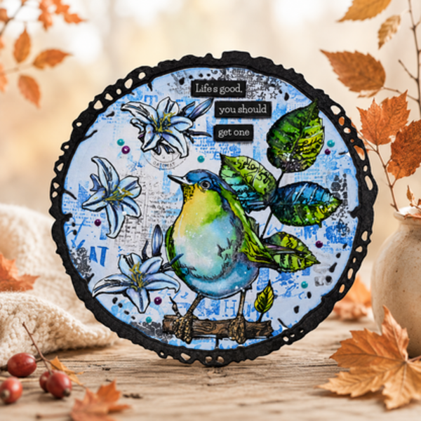 A circular mixed-media art print featuring a blue and green bird perched among white flowers and green leaves on a distressed blue background with the text 'Life's good, you should get one'. The print is displayed on a rustic wooden surface surrounded by autumn leaves, small red berries, and a soft beige fabric.