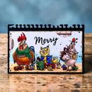 A rectangular printed card or art piece with a black spiral binding around the edges, featuring a colorful cartoon holiday scene with two roosters, presents, and the word “Merry” in script. It is photographed standing on a wooden surface against a soft blue blurred background.