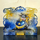 A decorative metal sign shaped like the zodiac sign Cancer, featuring a hand-painted cartoon cow holding a blue and gold egg. The sign has a distressed blue and yellow background with handwritten text and is displayed on a dark surface against a textured beige wall.