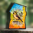 A handcrafted wooden plaque shaped like a house, featuring a colorful bird design with blue, yellow, orange, and green splashes, set against a blurred green background.