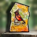 A house-shaped decorative plaque featuring a painted cardinal bird over a yellow, orange, and green distressed background with script and map textures, displayed on a wooden surface with a blurred green outdoor background.