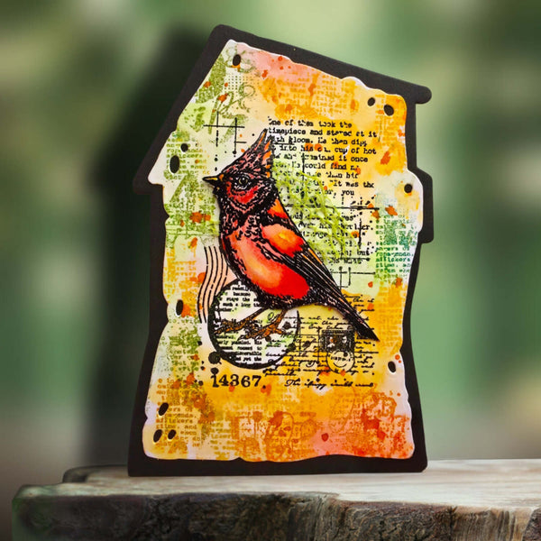 A house-shaped decorative plaque featuring a painted cardinal bird over a yellow, orange, and green distressed background with script and map textures, displayed on a wooden surface with a blurred green outdoor background.