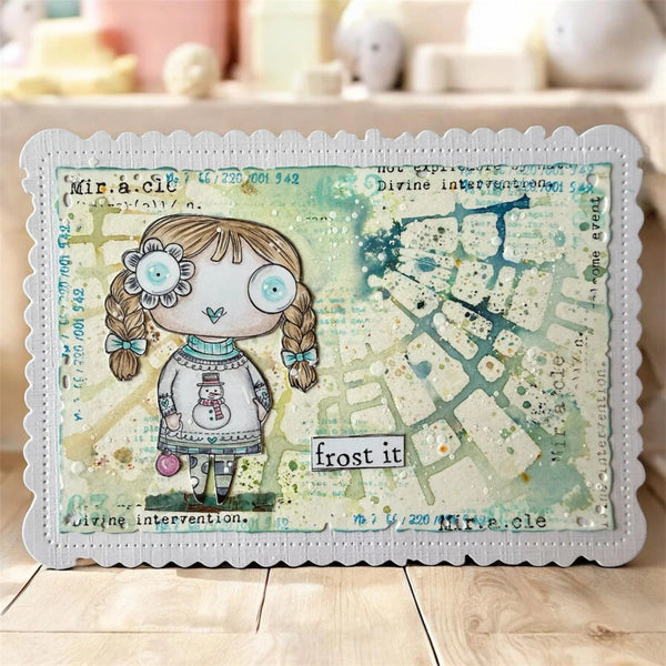 A rectangular mixed-media art card with scalloped edges, featuring a whimsical illustrated girl with braided pigtails, blue eyes, and a snowman sweater, alongside a distressed map design and text including 'frost it' and 'Miracle.' The card is photographed standing on a light wood surface with a softly blurred indoor background.