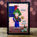 A rectangular greeting card with a black scalloped border, featuring a whimsical cartoon illustration of a person in a blue robe and Christmas tree hat holding a mug that says 'HALF DONE' and another card reading 'and fed up'. The card is displayed upright on a wooden surface against a dark background with warm bokeh lights.