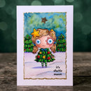A vertical greeting card featuring a whimsical watercolor illustration of a girl with blonde pigtails, blue eyes, and a flower in her hair, dressed in a Christmas tree outfit standing in front of Christmas trees. The card is displayed standing upright on a wooden surface with a blurred green bokeh background.