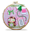A circular embroidery hoop containing a pink patterned fabric with green and pink thread, a cartoon girl holding a flower, the word "joy" in green script, and a small tag that reads "every stitch tells a story.".