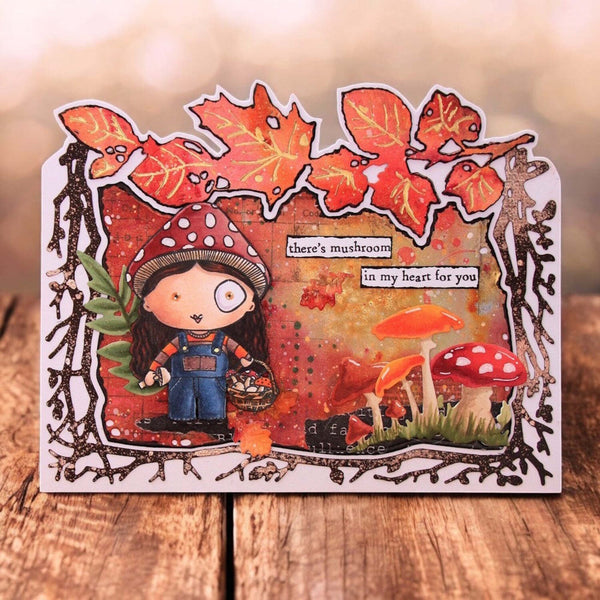 A handmade layered card featuring a whimsical cartoon girl in overalls under a red-and-white mushroom hat, surrounded by autumn leaves, mushrooms, and the text 'there's mushroom in my heart for you', displayed on a wooden surface.
