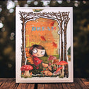 A square art card featuring a whimsical illustrated scene of a girl in a red dress sitting among mushrooms and trees, with the word “DREAM” in blue letters on an orange background, displayed on a wooden surface against a dark blurred backdrop.