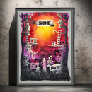 A framed abstract art print leaning against a textured concrete wall. The artwork features a stylized cityscape with layered black, white, and pink silhouettes, a bright orange and yellow center, and the word 'SHINE' on a distressed banner near the top.