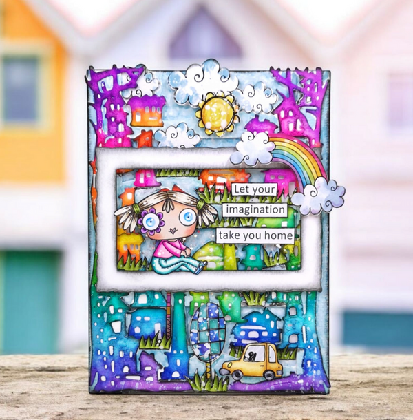 A colorful handmade picture frame card featuring a whimsical illustrated scene with a rainbow, clouds, sun, houses, trees, and a small character in the center. The card is displayed upright on a wooden surface with a blurred colorful house background.