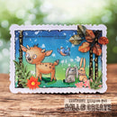 A handmade layered card featuring a cute woodland scene with a brown deer, a gray rabbit, a yellow bird, and autumn leaves on a blue sky background, displayed on a wooden surface with a blurred green background.