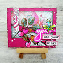 A handmade layered card displayed on a small wooden easel stand. The card features a bright pink border, a collage background with vintage maps and textures, a pink and green dragonfly, a pink heart-shaped bird, and layered text reading 'you use your wings'.
