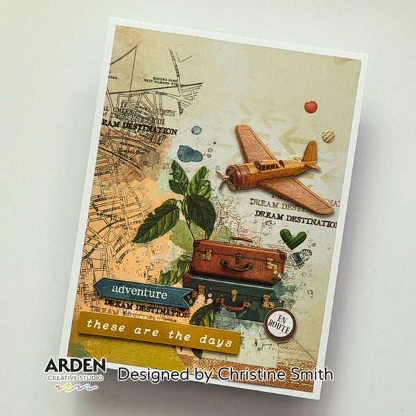 A handmade paper card featuring a vintage travel collage with a small wooden airplane, stacked suitcases, leaves, and text including 'DREAM DESTINATION' and 'ADVENTURE'. The card is photographed at an angle on a light gray background with the brand/designer text 'ARDEN' and 'Designed by Christine Smith' visible at the bottom.