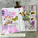An open mixed-media journal spread with purple, pink, white, and green floral and butterfly artwork, held open by a gold-toned metal clip on a gray textured background. Brand text at the bottom reads ARDEN and Designed by Katie Pertiet.