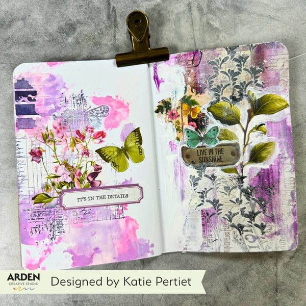 An open mixed-media journal spread with purple, pink, white, and green floral and butterfly artwork, held open by a gold-toned metal clip on a gray textured background. Brand text at the bottom reads ARDEN and Designed by Katie Pertiet.