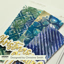 A close-up of a decorative handmade card or paper design featuring layered blue and green textured panels, embossed snowflake and tree motifs, and a gold script 'Christmas' element. A white banner at the bottom reads 'Designed by Christine Smith' with the Arden logo visible.