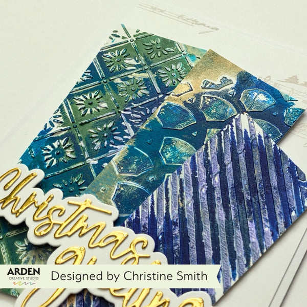 A close-up of a decorative handmade card or paper design featuring layered blue and green textured panels, embossed snowflake and tree motifs, and a gold script 'Christmas' element. A white banner at the bottom reads 'Designed by Christine Smith' with the Arden logo visible.