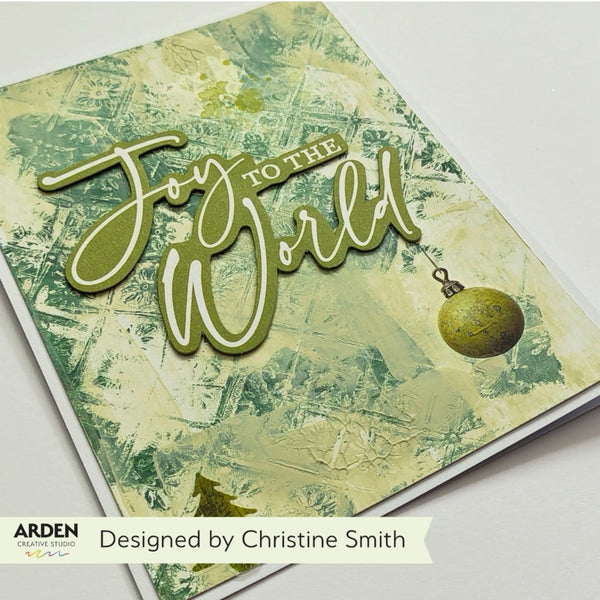 A handmade greeting card featuring the raised die-cut text 'Joy to the World' in olive green over a textured cream and green background, with a small ornament detail. The card is photographed on a white surface, with an ARDEN Creative Studios label and 'Designed by Christine Smith' text visible at the bottom.
