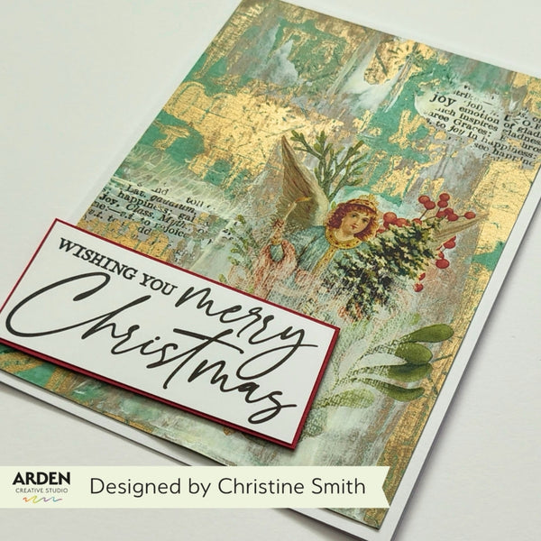 A handmade Christmas greeting card featuring a collage design with a gold-leaf textured background, vintage book text, a winged angel, red berries, and a white 'Wishing You Merry Christmas' banner. The card is photographed on a white surface with branding text 'ARDEN' and 'Designed by Christine Smith' at the bottom.