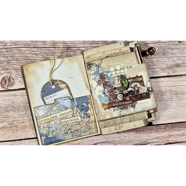 An open vintage-style collage journal or scrapbook album laid flat on a rustic wood plank surface. The pages are aged cream with layered collage elements including maps, postmarks, handwritten notes, small illustrations, and a hanging string tag.