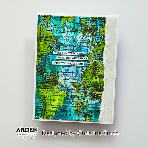 A handmade greeting card featuring a vintage-style world map design with green and blue watercolor washes, torn paper edges, and layered white text reading 'WISH YOU WERE HERE.' The card is photographed on a light gray background with Arden Creative Studio branding and 'Designed by Christine Smith' text below.