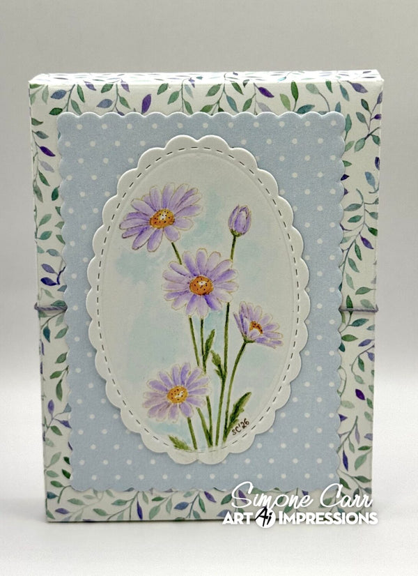 A handmade paper journal or scrapbook box featuring a light blue polka dot background with a central white scalloped oval containing painted purple daisies with yellow centers. The sides have a floral border pattern in green and purple.