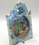 A decorative handmade gift box or favor box with a floral blue-and-white pattern, a light blue ribbon bow, and a hanging tag that reads 'home sweet home.' The front features a die-cut circular window showing a small illustrated house scene with a porch chair, flowers, and a birdhouse.