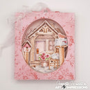 A pink floral square display frame with an oval cutout showing a whimsical house scene, a birdhouse, a mailbox with a bird, and flowers. A white ribbon bow is tied at the top, and the watermark 'Genette Houghton Art Impressions' is visible in the lower right corner.