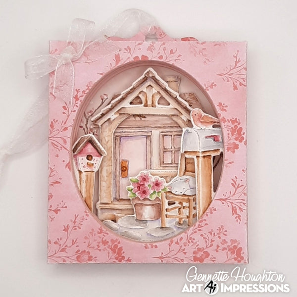 A pink floral square display frame with an oval cutout showing a whimsical house scene, a birdhouse, a mailbox with a bird, and flowers. A white ribbon bow is tied at the top, and the watermark 'Genette Houghton Art Impressions' is visible in the lower right corner.